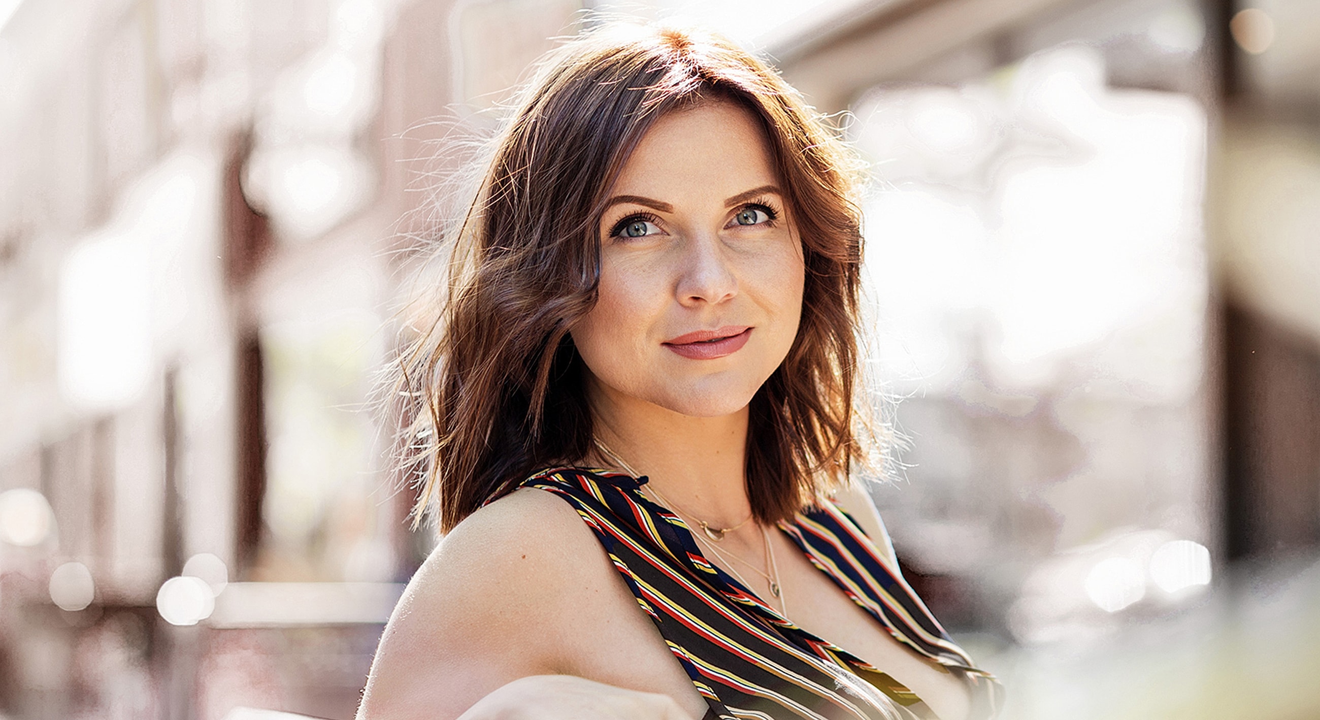 Woman smiling outdoors in sunny, urban setting.