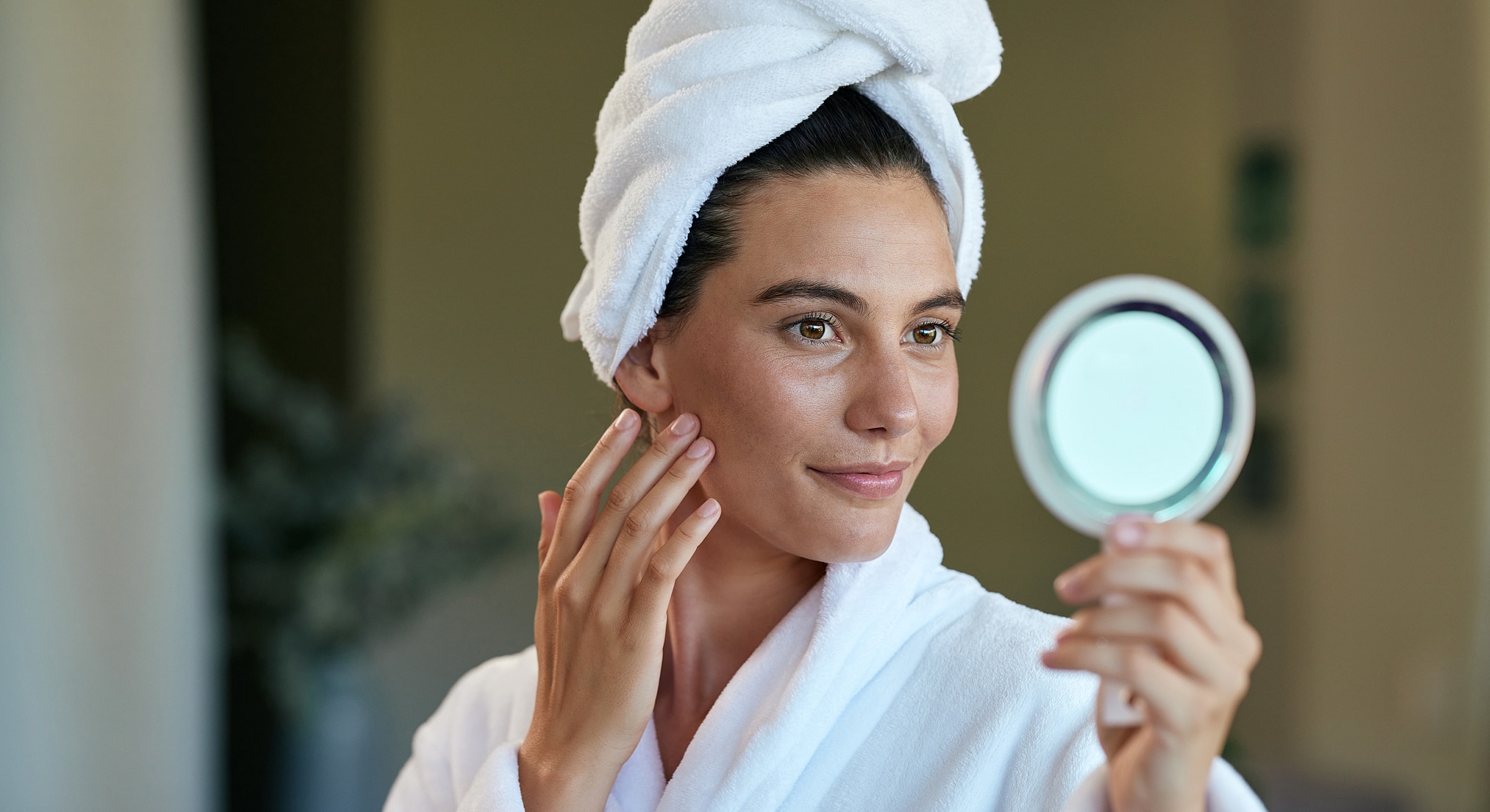 Woman with towel, applying skincare, looking in mirror.